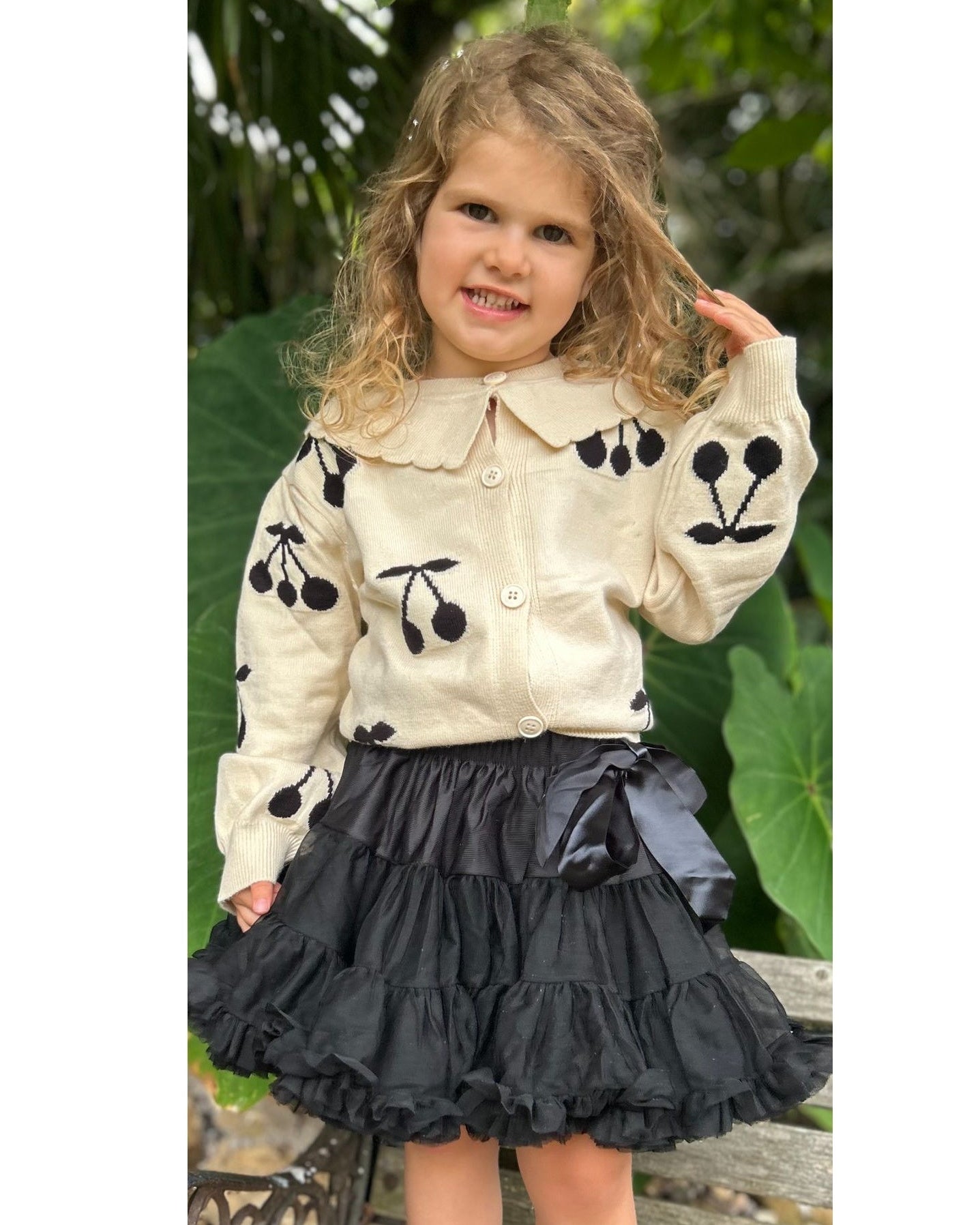 Young girl wearing a cream knitted cardigan with black cherry patterns and a black skirt outdoors.